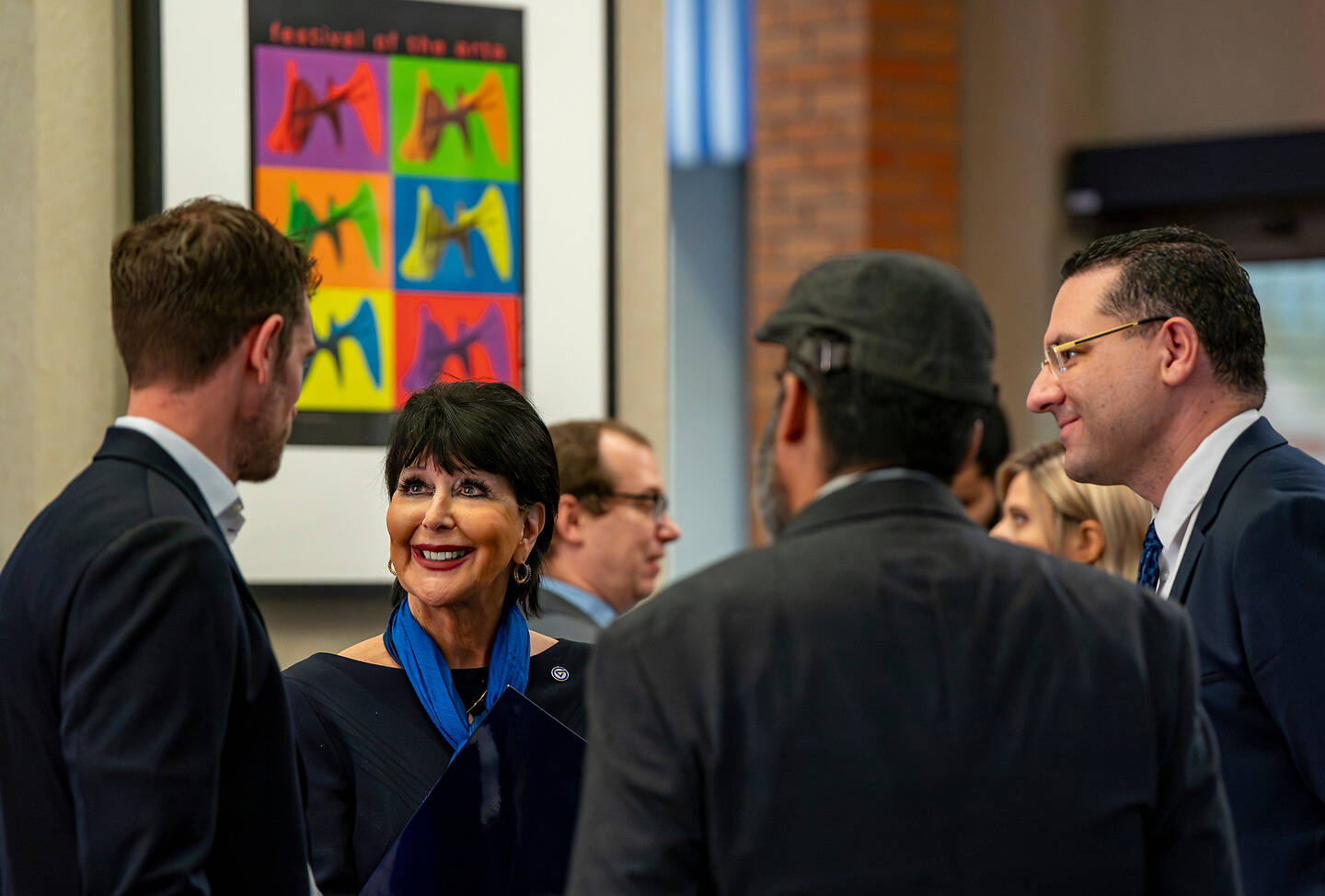 A group of faculty and GVSU President in formal attire engage in conversation at an indoor event, with a colorful art poster visible on the wall behind them.